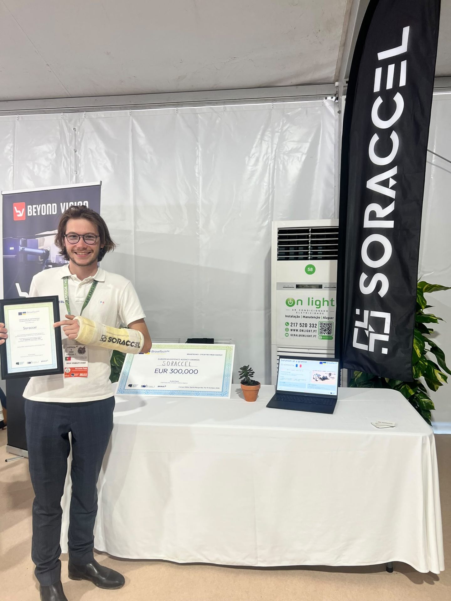 A man in a white polo shirt and a Soraccel-branded arm cast holds a framed certificate in front of the Soraccel stand, with the ceremonial EUR 300,000 award check on the table.